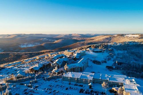 Aerial view of a snowy resort nestled in mountains, with buildings and trees blanketed in white under a clear blue sky.