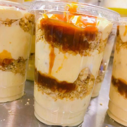 Layered dessert in a clear cup, featuring creamy filling, crumbled topping, and drizzled caramel sauce.