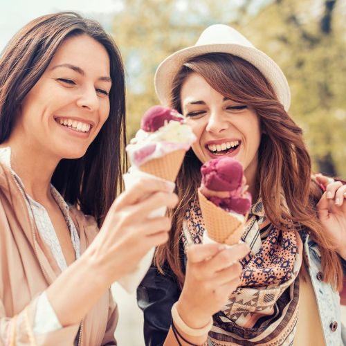 Two smiling women enjoying ice cream cones outdoors, wearing casual outfits and hats.