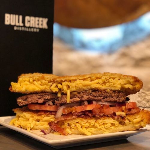 A stacked sandwich featuring a beef patty, bacon, tomatoes, and macaroni and cheese between two crispy layers.