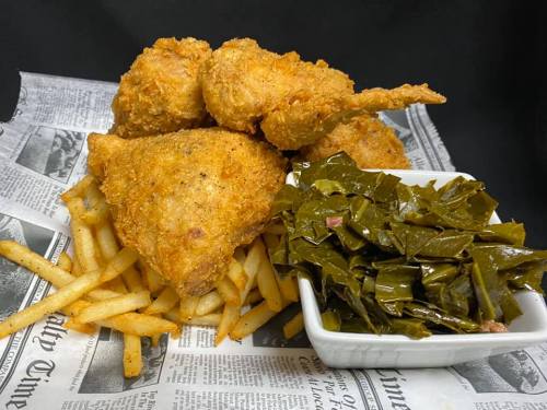 A plate of crispy fried chicken, golden fries, and a side of collard greens on a newspaper backdrop.