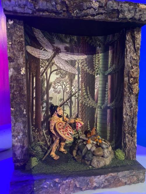 A diorama featuring a figure with a shield by a campfire in a forest setting, surrounded by trees and dragonflies.