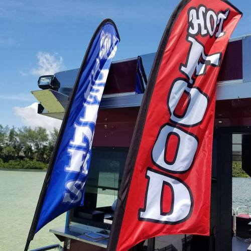 Two colorful flags on a boat: one blue with "Goodness" and one red with "Hot Food," set against a sunny sky.