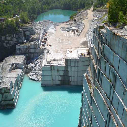 Aerial view of a quarry with turquoise water, stone blocks, and machinery surrounded by greenery.