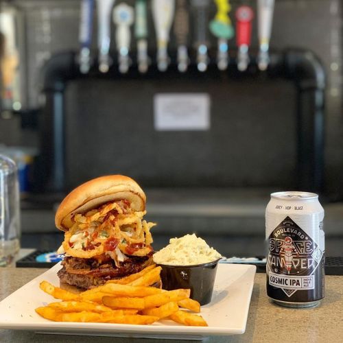 A hearty burger topped with bacon and onions, served with fries and coleslaw, alongside a can of Cosmic IPA.