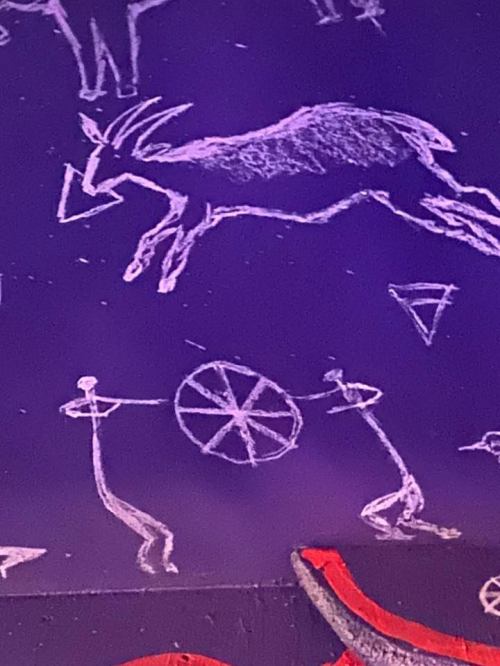 Ancient rock art featuring a goat and two figures holding a circular object, set against a dark background.