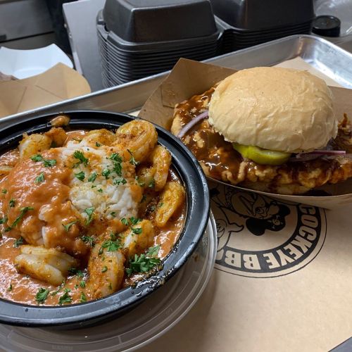 A bowl of shrimp with rice and sauce next to a BBQ sandwich with pickles and onions on a tray.