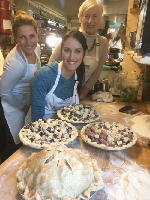 Three women in aprons smile behind freshly made fruit pies on a wooden table, with flour scattered around.