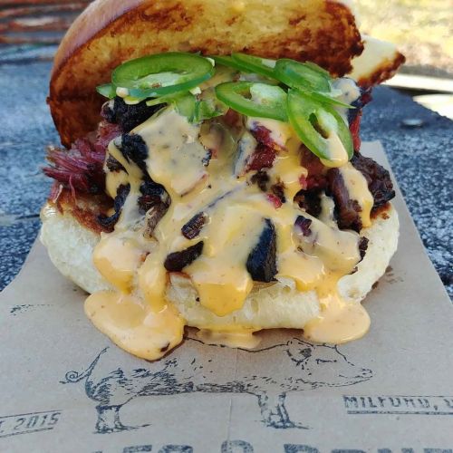 A close-up of a juicy burger with melted cheese, jalapeños, and smoky meat on a toasted bun, served on paper.