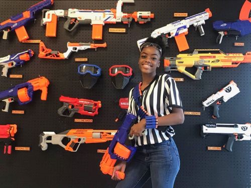 A person in a referee outfit poses with a toy blaster in front of a wall displaying various toy guns and accessories.