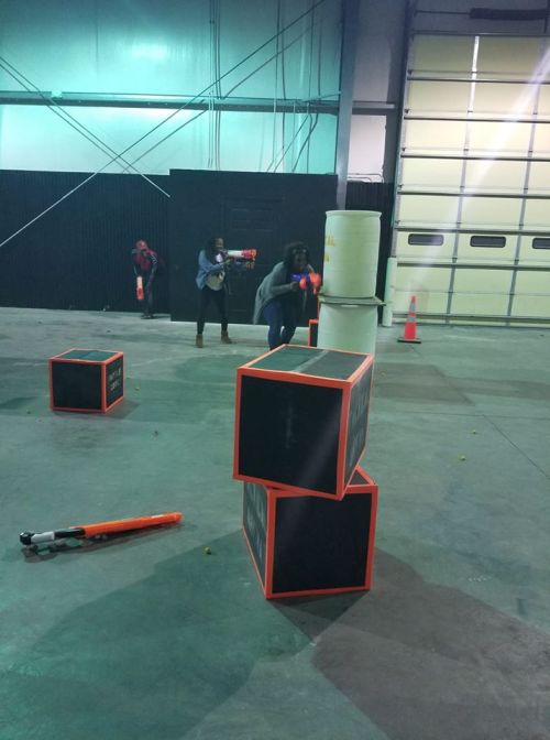 Two people play in an indoor arena, aiming toy blasters at targets behind stacked black boxes.