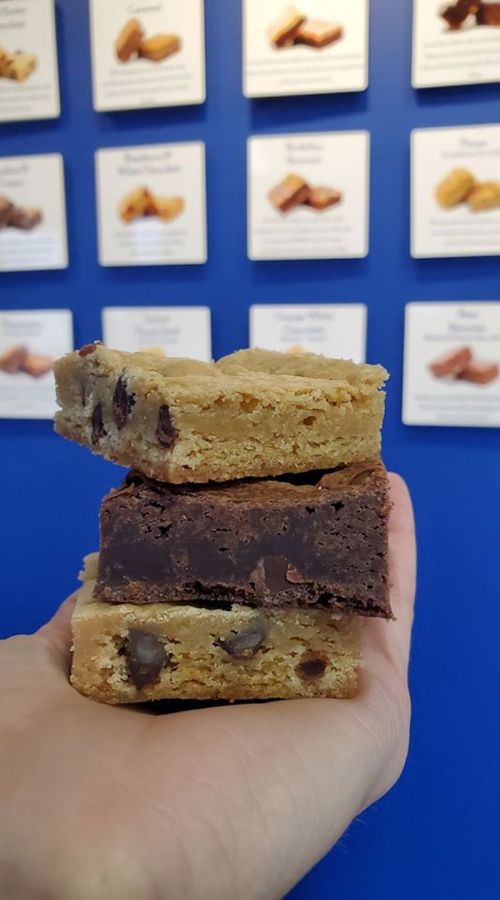 A hand holding three stacked dessert bars: a blondie on top, a brownie in the middle, and a chocolate chip bar at the bottom.