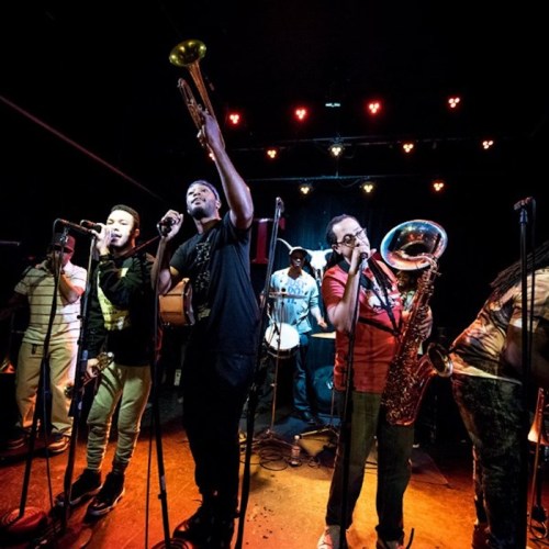 A lively band performs on stage, featuring musicians with various instruments and microphones under colorful lights.