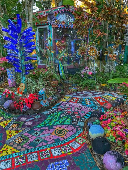 A vibrant garden path with colorful mosaics, plants, and a whimsical glass bottle tree leading to a decorated shed.
