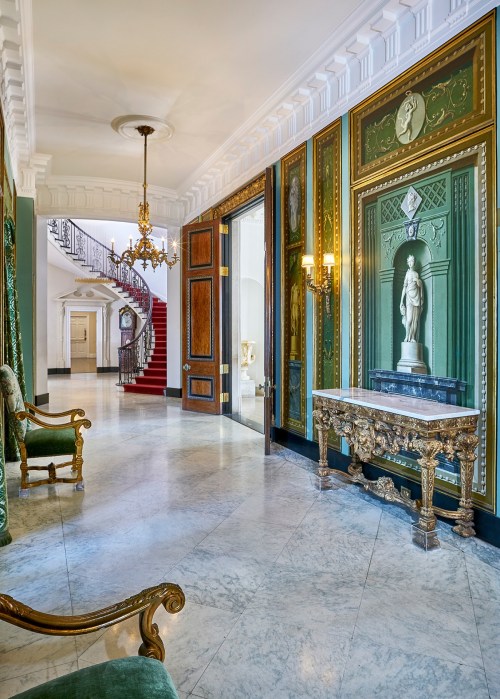 Elegant hallway with ornate decor, a statue, and a grand staircase in the background. Rich colors and intricate details.