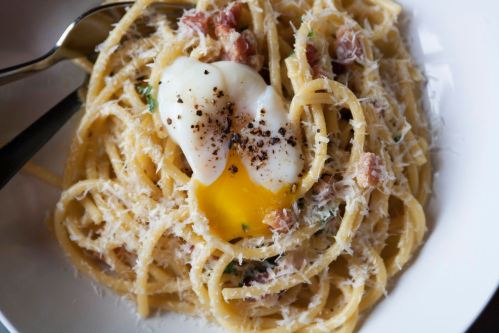 A bowl of spaghetti topped with a poached egg, crispy bacon, and grated cheese, garnished with black pepper.