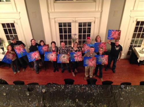 A group of people holding colorful paintings in a well-lit room with a long table and large windows.