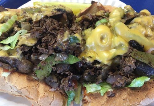 A loaded sandwich with seasoned beef, melted cheese, sautéed vegetables, and lettuce, served with a pickle on the side.