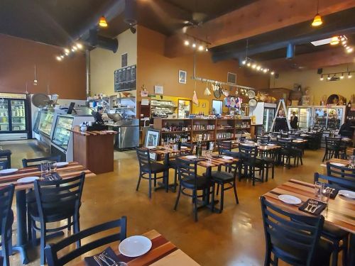 Interior of a restaurant featuring wooden tables, chairs, and shelves with food items, with a staff member in the background.