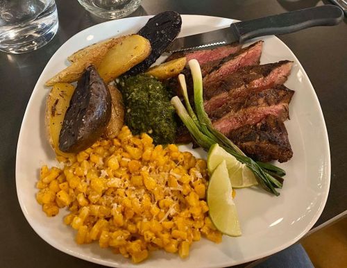 A plate of grilled steak slices, roasted potatoes, corn, green sauce, and lime wedges, garnished with green onions.