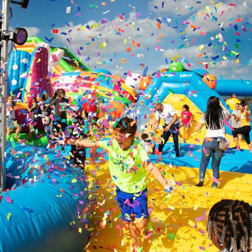 A lively scene of children playing on inflatable structures, surrounded by colorful confetti and a bright blue sky.