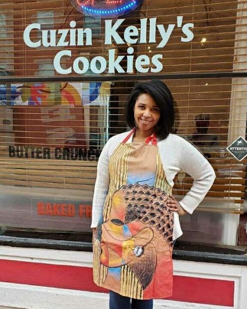 A woman in an artistic apron stands in front of a cookie shop window, smiling confidently.