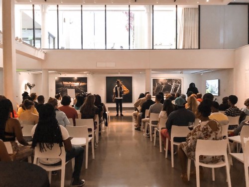 A speaker addresses a seated audience in a modern gallery with large windows and artwork on the walls.