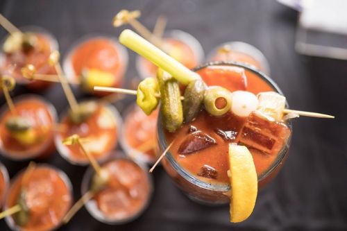 A close-up of a cocktail garnished with celery, olives, pickles, and lemon, with several similar drinks in the background.
