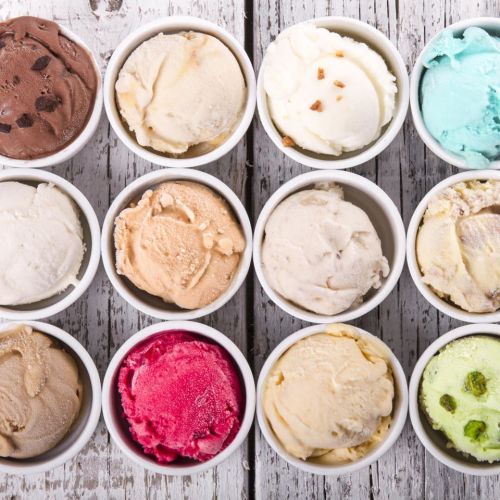 Twelve small bowls of various ice cream flavors arranged on a rustic wooden surface.