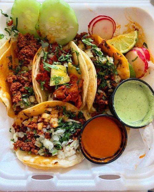 Three tacos filled with meat, onions, and cilantro, served with lime, cucumber, radishes, and two sauces in containers.