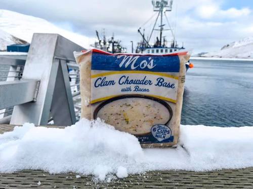 A package of Mo's Clam Chowder Base with Bacon sits on snow near a harbor with fishing boats in the background.