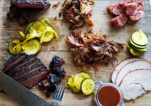 A wooden board with various meats: brisket, pulled pork, sausage, and turkey, accompanied by pickles and barbecue sauce.