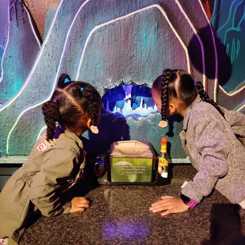 Two children with braided hair lean over a table, peering into a colorful cave mural.