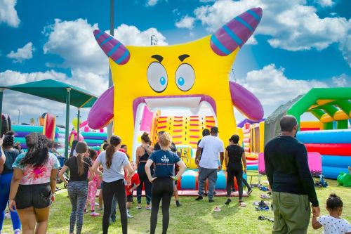 A colorful inflatable slide with a monster face, surrounded by children and adults enjoying a sunny day at a park.