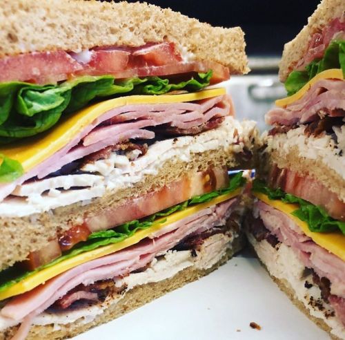 Close-up of a layered sandwich with turkey, ham, cheese, lettuce, and tomato on whole grain bread.