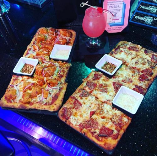 Two rectangular pizzas on a black countertop, one topped with pepperoni and the other with vegetables, alongside dipping sauces.