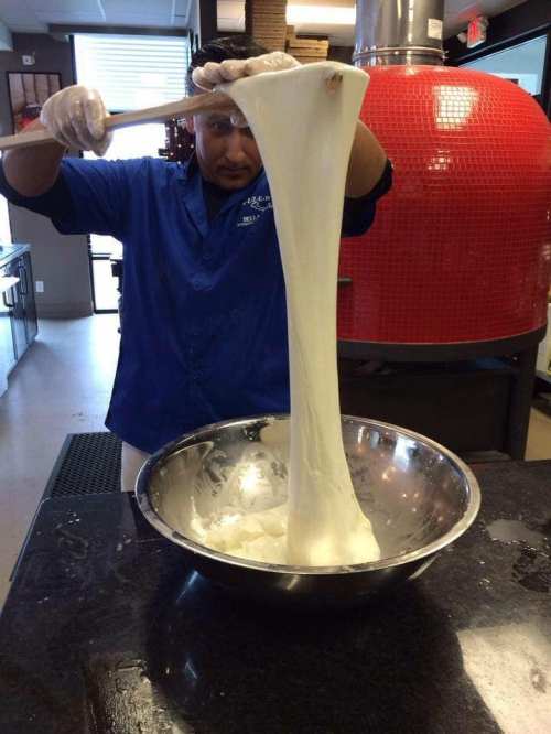 A chef in a blue jacket stretches a large batch of dough over a mixing bowl in a kitchen setting.