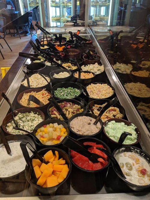 A buffet display featuring various bowls of colorful salads, fruits, and pasta, each with a serving spoon.