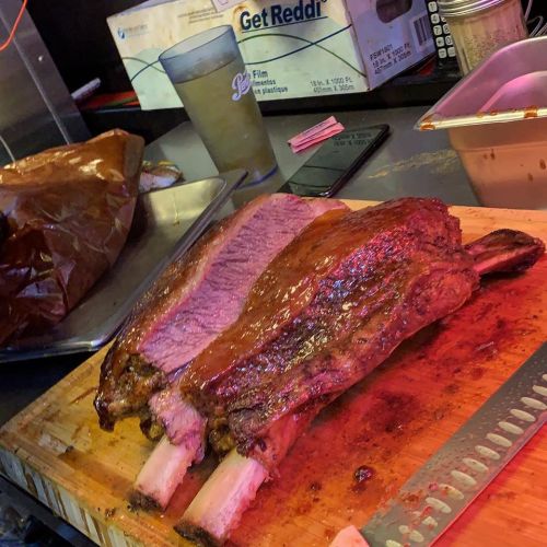 A large, juicy rack of ribs on a wooden cutting board, with a knife and other kitchen items in the background.