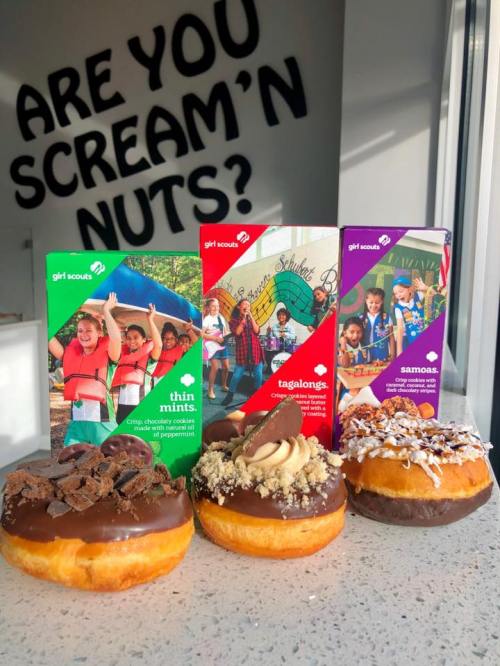 Three donuts topped with chocolate and various toppings, with Girl Scout cookie boxes in the background.