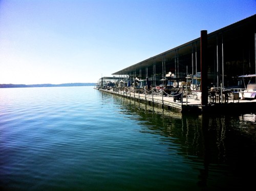 A calm lake scene with a wooden dock and a modern building reflecting on the water under a clear blue sky.