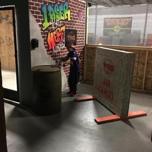 A child plays in an indoor Nerf battlefield, surrounded by colorful graffiti and obstacles like barrels and walls.