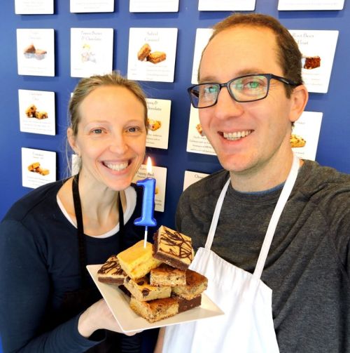 A smiling couple holds a plate of desserts with a birthday candle, celebrating a milestone in front of a dessert display.