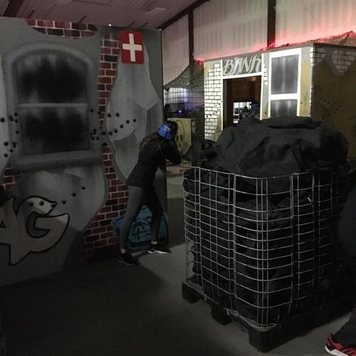 A person in a dark room aims a toy gun, surrounded by graffiti-covered walls and a large black container.