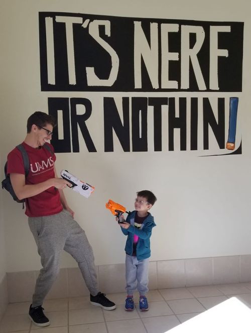 A young boy playfully aims a Nerf gun at a teenager, who holds a Nerf blaster, in front of a wall with "IT'S NERF OR NOTHIN!"