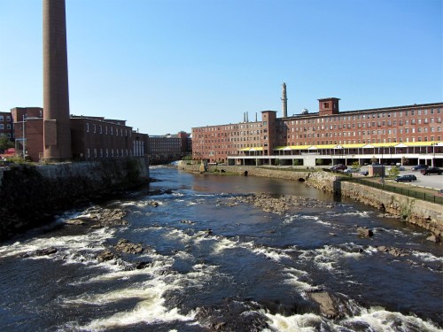 A river flows past historic brick buildings and a tall smokestack under a clear blue sky.