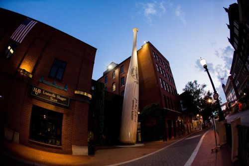 A large baseball bat sculpture stands outside the Louisville Slugger Store at dusk, with buildings and streetlights nearby.