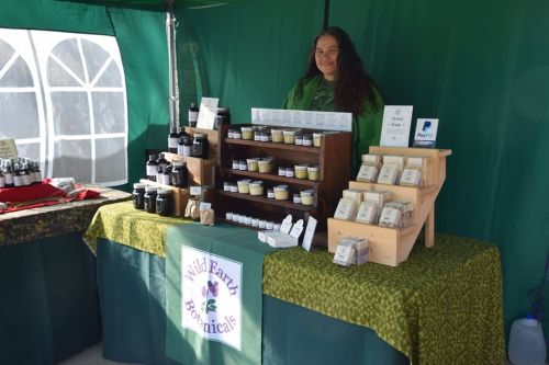 A vendor stands behind a table displaying various natural products at a market, with a green tent backdrop.