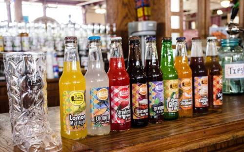 A row of colorful soda bottles on a wooden counter, with a glass and tips jar in the background.