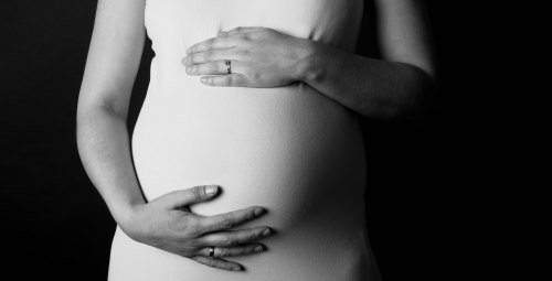 A close-up of a pregnant person's hands gently resting on their belly, set against a dark background.